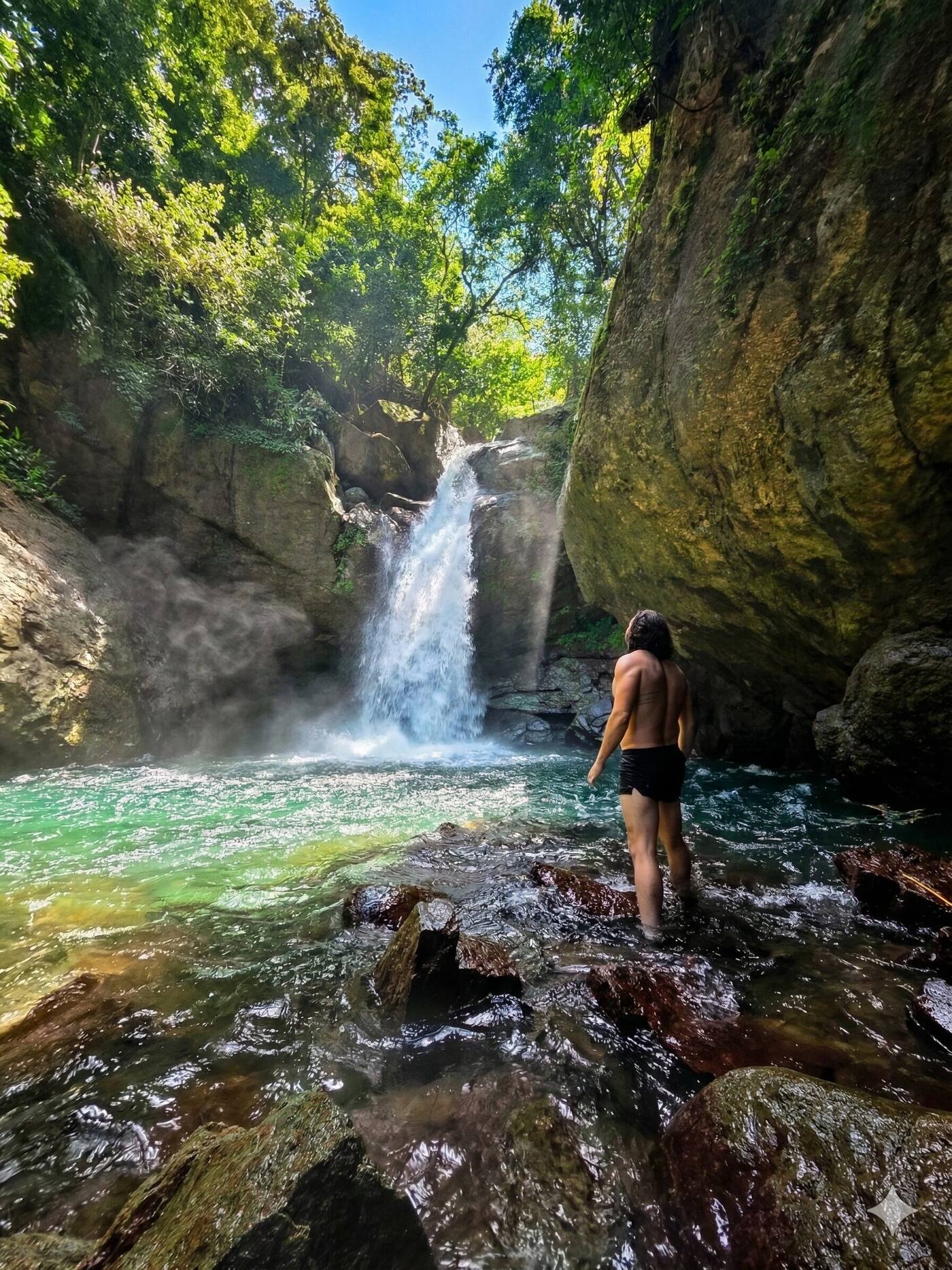 A guest stands in a turquoise jungle river pool, facing a waterfall surrounded by green canopy.